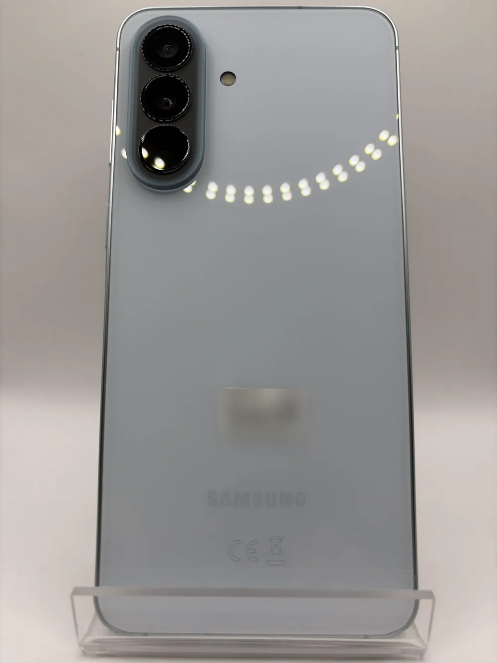 Samsung Galaxy A57 5G gris clair, vu de dos, triple capteur photo, posé sur un support transparent