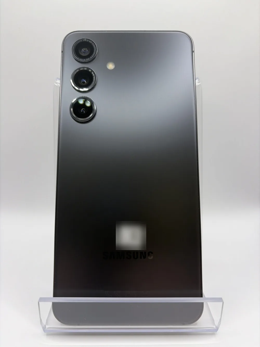 Samsung Galaxy S24 gris anthracite, vu de dos, trois capteurs photo alignés, posé sur un support transparent